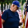 A senior veteran in a blue cap stands beside American flags, honoring service.
