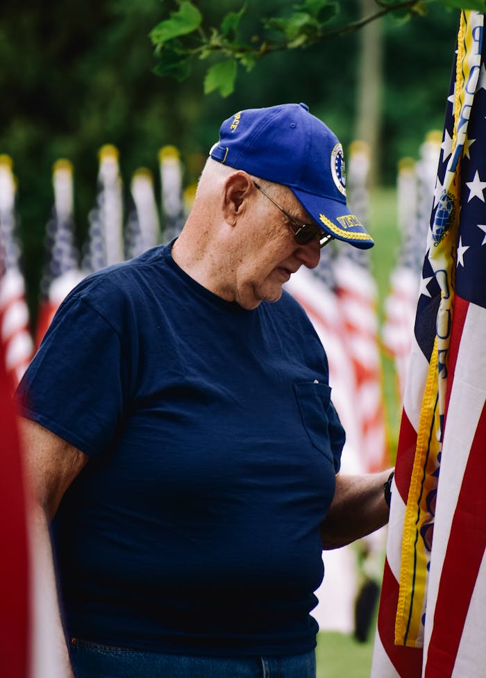 A senior veteran in a blue cap stands beside American flags, honoring service.